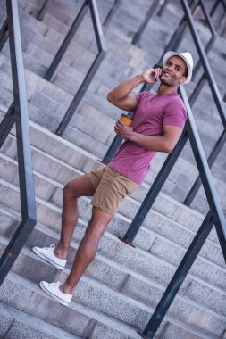 Yakışıklı adam cep telefonuyla konuşuyor, elinde bir fincan kahve tutuyor ve merdivenlerde dikilirken gülümsüyor.