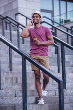 Yakışıklı adam cep telefonuyla konuşuyor, elinde bir fincan kahve tutuyor ve şehirde yürürken gülümsüyor.