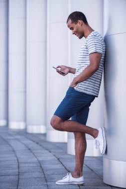 Yakışıklı bir adamın akıllı bir telefon kullanıp şehirde yürürken gülümsemesinin tam boy portresi.