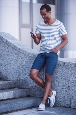 Yakışıklı bir adamın akıllı bir telefon kullanıp şehirde yürürken gülümsemesinin tam boy portresi.