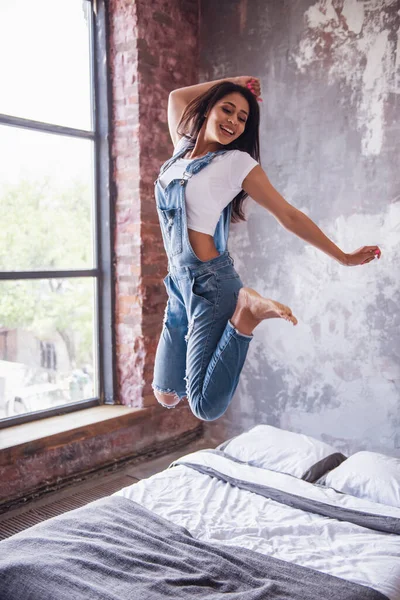 Beautiful girl in denim overall is looking at camera and smiling while jumping on bed at home