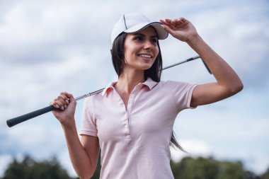 Güzel genç kadın bir golf kulübü tutarak ve golf sahası üzerinde dururken gülümseyen