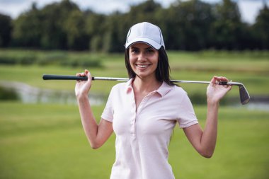 Güzel genç kadın bir golf kulübü holding, kameraya bakıyor ve golf sahası üzerinde dururken gülümseyen