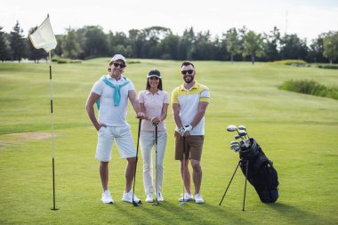 İki erkek ve bir kadın golf kulüpleri holding, kameraya bakıyor ve golf sahası üzerinde dururken gülümseyen