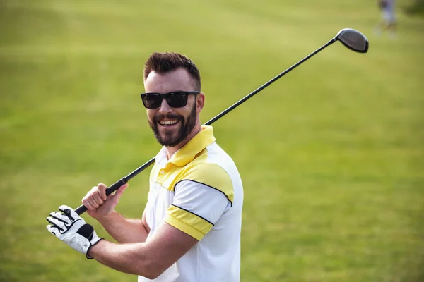 Handsome guy is holding a golf club, looking at camera and smiling while standing on golf course