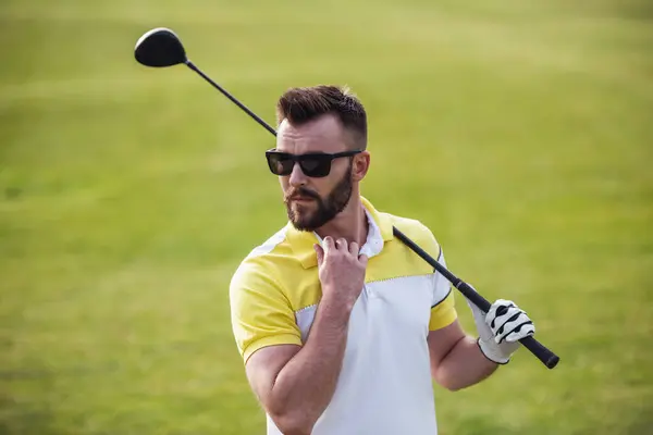 Handsome guy is holding a golf club and looking away while standing on golf course