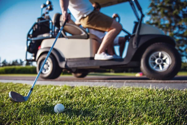 Cropped image of handsome men driving a golf cart, one guy is raising a golf club at the ball, ball in focus