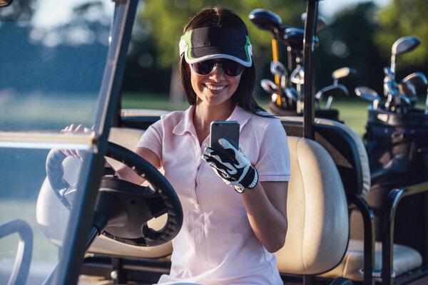 Beautiful young woman is using a smart phone and smiling while driving a golf cart