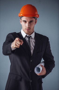 Takım elbise ve koruyucu kask yakışıklı genç mimar taslak tutarak ve kamera, gri arka plan üzerine işaret