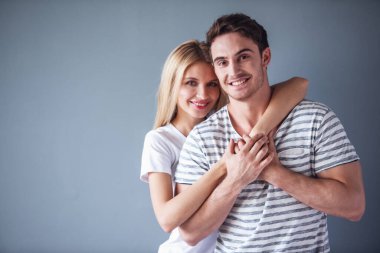 Güzel genç çift sarılma, kameraya bakıyor ve gülümsüyor, gri arka plan üzerinde