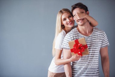 Güzel genç kadın bir hediye kutusu tutuyor ve bir sürpriz yapma erkek gözlerini kapsayan, her ikisi de, gri arka plan üzerinde gülümsüyor musun