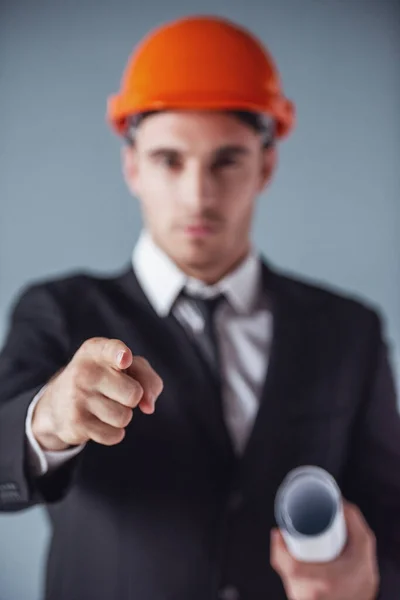 Handsome young architect in suit and protective helmet is holding draft and pointing at camera, on gray background, hand in focus