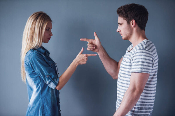 Beautiful young man and woman are pointing hand-guns at each other, on gray background