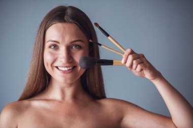 Güzel kadın çıplak omuz ile makyaj fırçaları, kameraya bakıyor ve gülümsüyor, gri arka plan üzerinde tutuyor