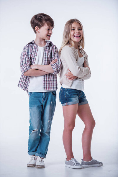 Full length of beautiful little girl and boy looking at camera and laughing while standing with crossed arms on light background