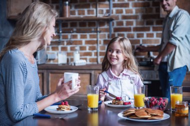 Mutlu bir aile mutfakta kahvaltı. Baba krep yaparken anne ve kızı masada konuşuyor