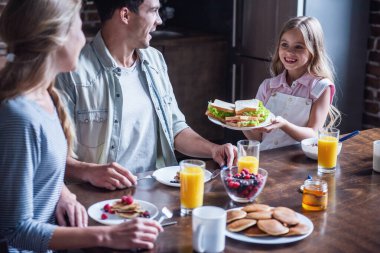 Mutlu bir aile mutfakta kahvaltı. Küçük kızın ailesine sandviç getiren ve gülümseyen