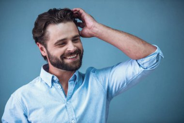 Yakışıklı sakallı iş adamı akıllı rahat kıyafet saçları düzgünleştirme ve gri arka plan üzerinde gülümseyen