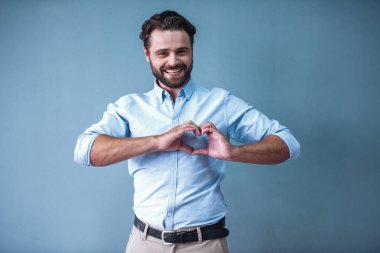 Yakışıklı sakallı iş adamı akıllı rahat kıyafet bir kalp, kameraya bakıyor ve gülümsüyor, gri arka plan üzerinde gösterilen