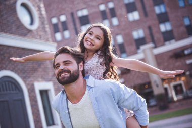 Yakışıklı sakallı baba ve sevimli küçük kızını birlikte dışarıda oynarken gülümsüyor musun, kız sırtıma oturuyor