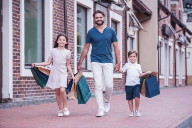 Yakışıklı sakallı baba ve sevimli küçük çocuklarını alışveriş çantaları taşırken, el ele tutuşarak ve sokakta yürürken gülümseyen