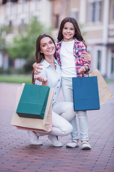 Beautiful mom and her cute little daughter are holding shopping bags, looking at camera and smiling while standing outdoors