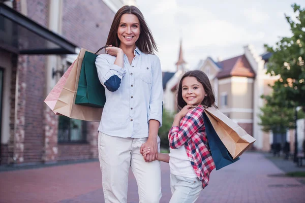 Beautiful mom and her cute little daughter are carrying shopping bags, holding hands, looking at camera and smiling while standing outdoors