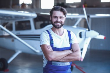 Üniformalı yakışıklı sakallı makinist kameraya bakıyor ve hangarda uçak yakın çapraz kolları olan dururken gülümseyen
