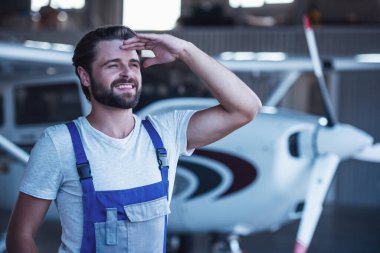 Üniformalı yakışıklı sakallı makinist uzak ve gülümseyen yakınındaki bir uçak hangarda dururken arıyor