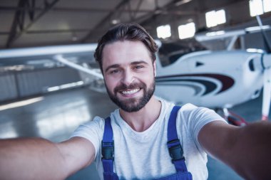 Üniformalı yakışıklı sakallı makinist selfie yapıyor ve yakınlarında bir uçak hangarda dururken gülümseyen