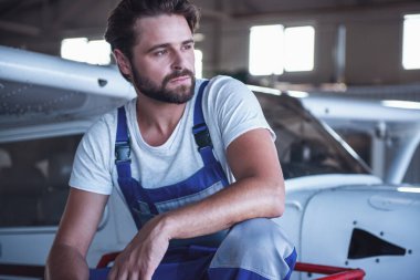 Üniformalı yakışıklı sakallı makinist uzakta otururken hangarda uçak yakınındaki arıyor