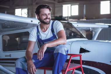 Üniformalı yakışıklı sakallı mekanik Dış Saha ve gülümseyen hangarda uçak yakınındaki otururken arıyor