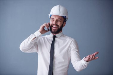 Yakışıklı genç işadamı elbiseli sakallı ve koruyucu kask cep telefonda konuşurken ve gri arka plan üzerinde gülümseyen