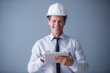 Takım elbise ve koruyucu kask yakışıklı olgun işadamı dijital tablet, kameraya bakıyor ve gülümsüyor, gri arka plan üzerinde kullanma
