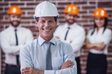 Takım elbise ve koruyucu kask yakışıklı olgun işadamı kameraya bakıyor ve gülümsüyor, arkadaşları içinde belgili tanımlık geçmiş