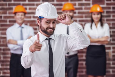 Takım elbise ve koruyucu kask yakışıklı sakallı işadamı kameraya bakıyor ve gülümsüyor, arkadaşları içinde belgili tanımlık geçmiş