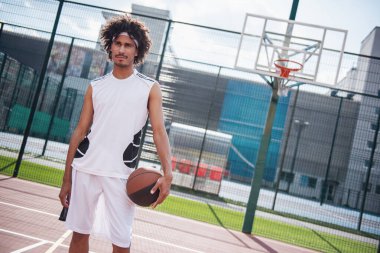 Çekici basketbol oyuncusu topu tutuyor, uzağa bakıyor ve dışarıda basketbol sahasında dururken gülümsüyor.