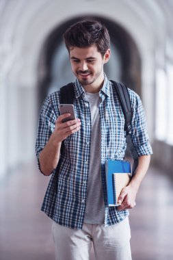 Yakışıklı genç öğrenci bir akıllı telefon kullanarak ve üniversite salonunda dururken gülümseyen