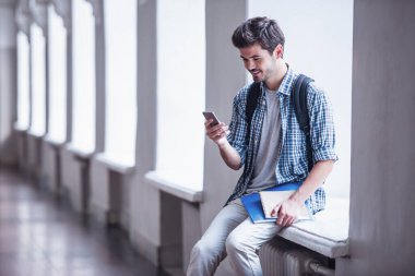Yakışıklı genç öğrenci bir akıllı telefon kullanarak ve üniversite salonunda pencere eşiğinde otururken gülümseyen