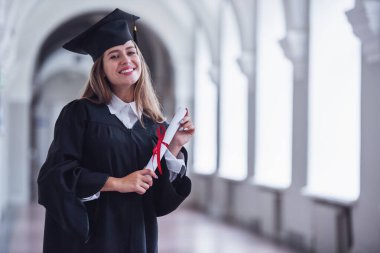 Akademik elbiseli çekici genç kadın yüksek lisans diplomasına tutarak, kameraya bakıyor ve Üniversitesi Salonu'nda dururken gülümseyen