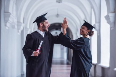 Akademik elbiseler mutlu genç mezunlar diploma holding, çak bir beşlik veren ve üniversite salonunda dururken gülümseyen
