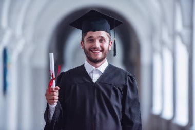 Akademik elbiseli yakışıklı genç lisansüstü diploma holding, kameraya bakıyor ve Üniversitesi Salonu'nda dururken gülümseyen