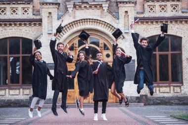 Akademik elbiseler başarılı mezunlar diploma holding, kameraya bakıyor ve fotoğraf için açık havada atlama sırasında gülümseyen
