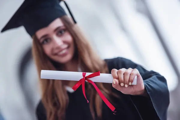 Attractive young female graduate in academic dress is holding a diploma, looking at camera and smiling while standing in university hall