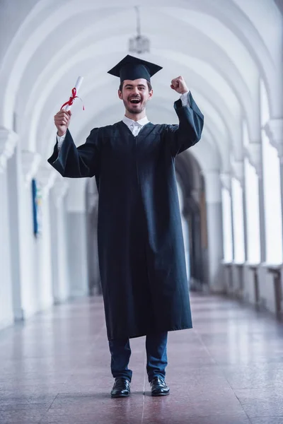 Handsome  young graduate in academic dress is holding diploma, looking at camera and smiling while standing in university hall
