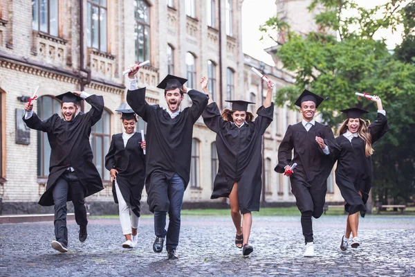 Successful graduates in academic dresses are holding diplomas, looking at camera and smiling while running outdoors