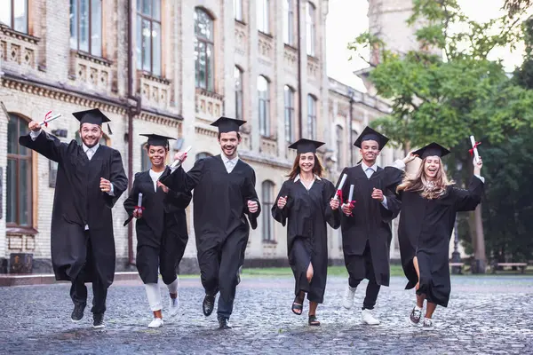 Successful graduates in academic dresses are holding diplomas, looking at camera and smiling while running outdoors