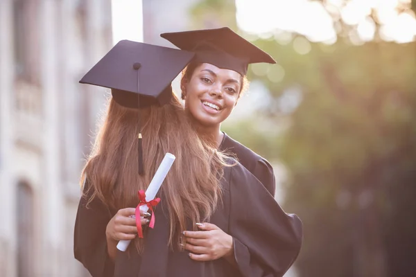 Beautiful female graduates in academic dresses are hugging and smiling while standing outdoors
