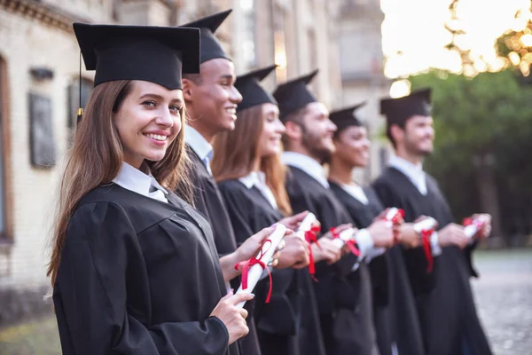 Successful graduates in academic dresses are holding diplomas, looking forward and smiling while standing in a row outdoors