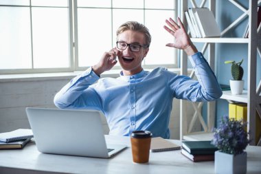 Resmi kıyafetler yakışıklı genç işadamı cep telefonda konuşurken, bir dizüstü bilgisayar kullanarak ve ofiste çalışırken gülümseyen